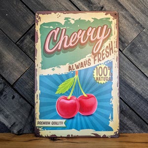 May include: A vintage-style metal sign with the word "Cherry" in a pink and brown font. The sign features two red cherries with green leaves, the text "Always Fresh," and "100% Natural."