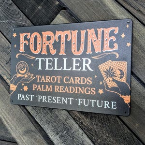 May include: A rectangular black metal sign with peach and white text and graphics. It reads FORTUNE TELLER, TAROT CARDS, PALM READINGS, and PAST PRESENT FUTURE. Illustrations include a hand holding a crystal ball and a hand holding tarot cards.