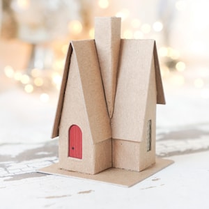 Puede incluir: Una pequeña casa de cartón con una puerta roja y una chimenea. La casa está hecha de cartón de color beige y se asienta sobre una base rectangular. El fondo está borroso con luces suaves y desenfocadas.