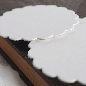 May include: Two white scalloped paper circles, stacked on top of each other, on a brown textured surface.