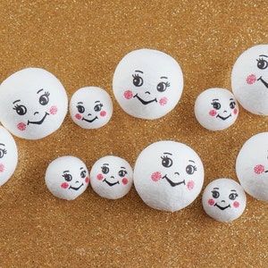 Puede incluir: Una colección de bolas de espuma blanca con caras pintadas. Las bolas son de varios tamaños y tienen ojos negros, bocas negras y mejillas rosadas.