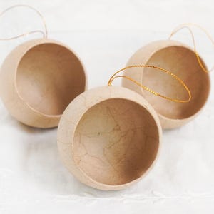 May include: Three spherical, open-faced ornaments with a natural, light brown paper texture. Each ornament has a gold-colored string for hanging. The ornaments are arranged on a white, textured surface, suggesting a craft or holiday theme.