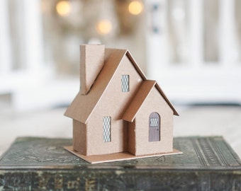 May include: A small cardboard model of a house with a chimney, windows, and a door. The house is sitting on top of an old book.