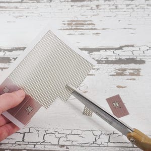 May include: A person is cutting a sheet of paper with a pair of scissors. The paper has a grid pattern and two small rectangular shapes with a window cut out. The paper is on a white wooden surface.
