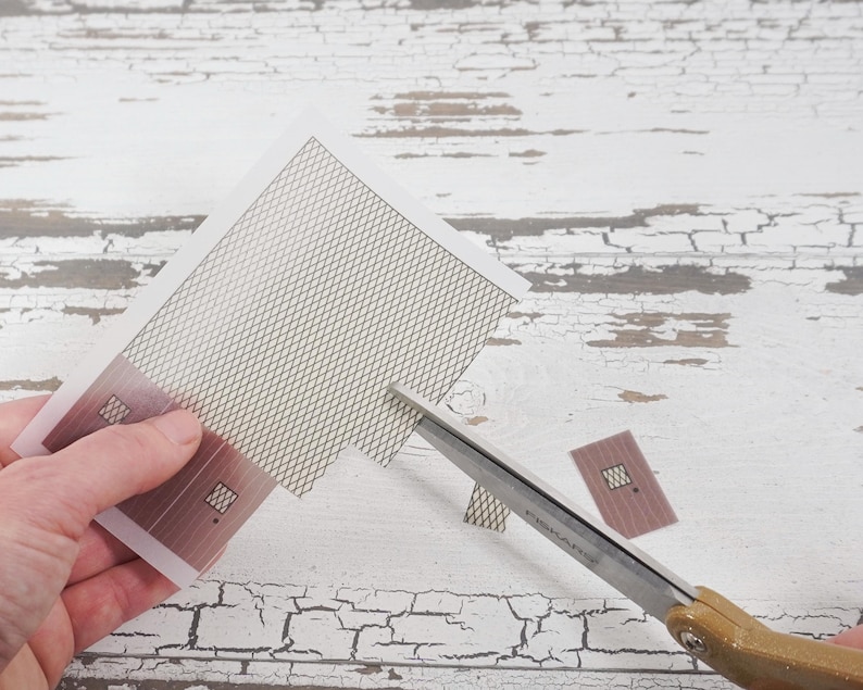 May include: A person is cutting a sheet of paper with a pair of scissors. The paper has a grid pattern and two small rectangular shapes with a window cut out. The paper is on a white wooden surface.