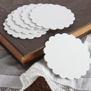 May include: A stack of six white scalloped paper circles, with one circle laying flat on a white lace tablecloth. The circles are blank and can be used for crafts, gift tags, or other projects.