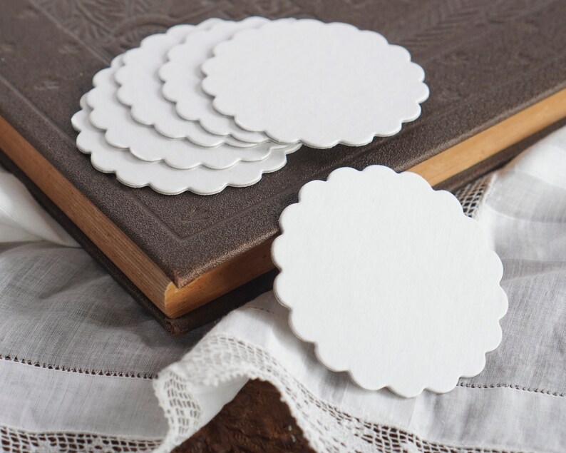 May include: A stack of six white scalloped paper circles, with one circle laying flat on a white lace tablecloth. The circles are blank and can be used for crafts, gift tags, or other projects.