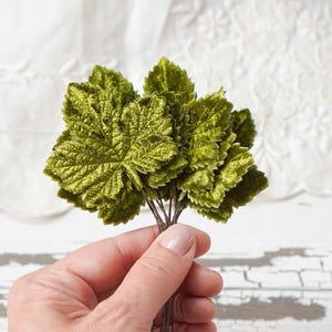 May include: A cluster of artificial green velvet leaves, each with detailed veining, held together by thin brown stems. The leaves have a textured, plush appearance, and the overall effect is a vibrant, naturalistic floral element.