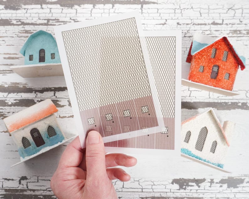 May include: A hand holds two sheets of paper with a grid pattern. The paper is white with a brown grid pattern. The paper is being held over a white wooden surface with miniature houses on it. The houses are blue, orange, and white.