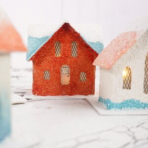 May include: Three miniature paper houses with white, red, and blue paint. The houses have windows and doors, and the roofs are covered in white snow.