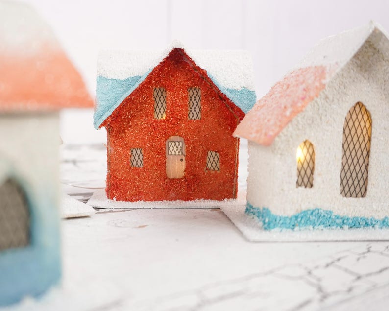 May include: Three miniature paper houses with white, red, and blue paint. The houses have windows and doors, and the roofs are covered in white snow.