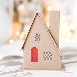 Puede incluir: Una pequeña casa de cartón marrón con una puerta roja y dos ventanas enrejadas. La casa tiene una chimenea y está colocada sobre una superficie de madera blanca.