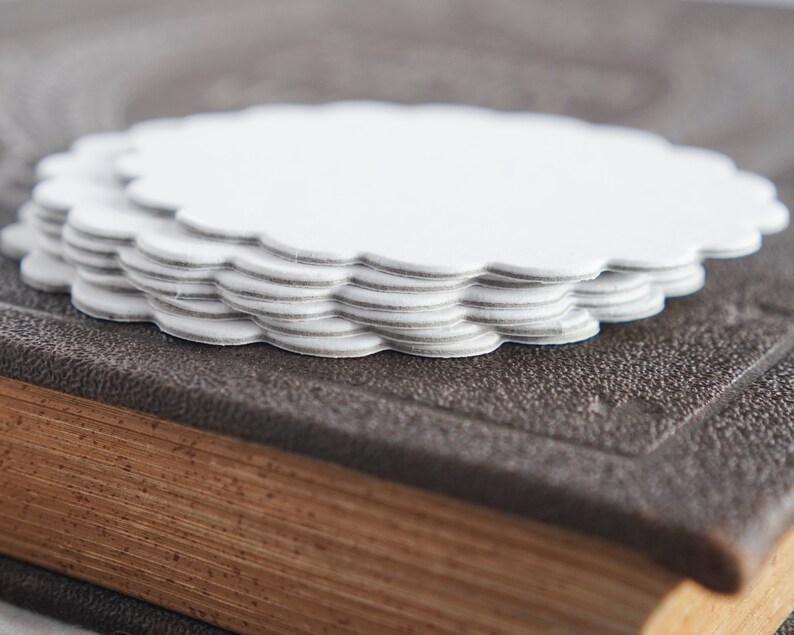 May include: A stack of white paper doilies with scalloped edges, sitting on top of a brown leather-bound book.