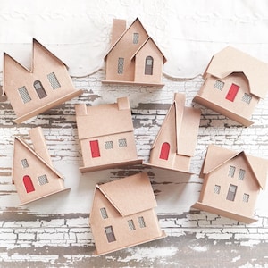 Puede incluir: Una colección de casas de cartón en miniatura de varios diseños. Cada casa presenta un exterior de color beige, con detalles como puertas rojas o marrones y ventanas con patrones. Algunas tienen chimeneas o tejados inclinados, creando una escena encantadora.