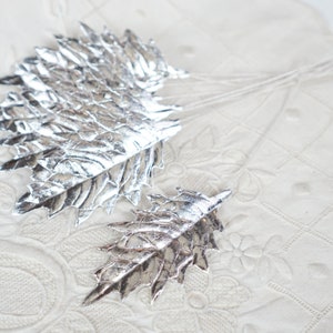 Op de afbeelding: Twee zilverkleurige bladvormige decoraties met gedetailleerde nerven. Ze liggen op een witte, getextureerde ondergrond. De bladeren hebben een metallic glans en zijn waarschijnlijk gemaakt van metaal of een vergelijkbaar materiaal. De afbeelding suggereert een decoratief of ambachtelijk thema.