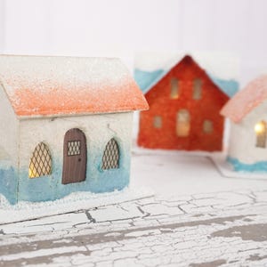 May include: Three miniature paper houses with white, red, and blue paint. The houses have a snowy roof and windows with lights inside. The houses are sitting on a white, cracked surface.