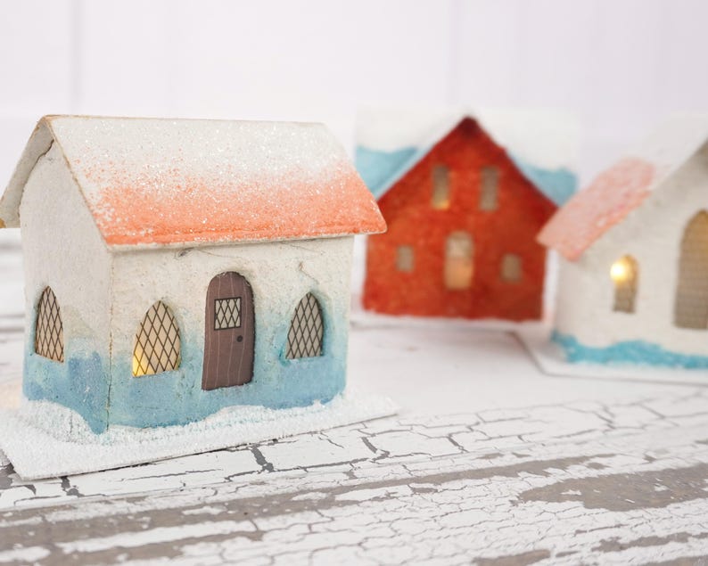May include: Three miniature paper houses with white, red, and blue paint. The houses have a snowy roof and windows with lights inside. The houses are sitting on a white, cracked surface.