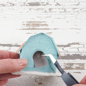 May include: A light blue and orange miniature house with a white base. The house has a small window with a grid pattern. A person is using tweezers to place the window in the house.