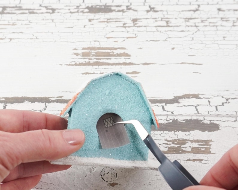 May include: A light blue and orange miniature house with a white base. The house has a small window with a grid pattern. A person is using tweezers to place the window in the house.