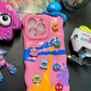 Puede incluir: Una funda de teléfono rosa con un diseño azul, amarillo y morado, adornada con figuras de calamares de dibujos animados de colores. Una figura azul con forma de pulpo se encuentra en la parte superior. La funda tiene recortes para la cámara y los botones.