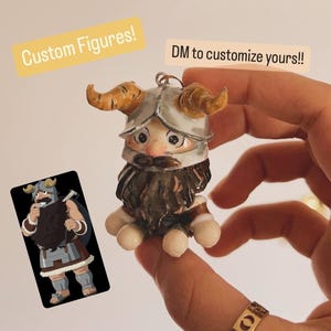 May include: A handcrafted miniature Viking figure with a horned helmet, a long brown beard, and a mustache. The figure is holding a small card with a cartoon Viking illustration. Text on the image reads "Custom Figures!" and "DM to customize yours!!"