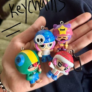 May include: Four character keyrings in various colours. The keyrings depict cartoon figures with different designs, including a skull, a star, and a Santa hat. The word "keychains" is written in black marker above the keyrings.