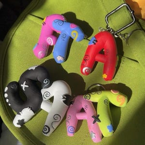 May include: A selection of handmade letter keyrings in the shape of the letter "A". The keyrings are painted in vibrant colours such as pink, blue, red, black, white, and green. Each letter features unique decorative designs.