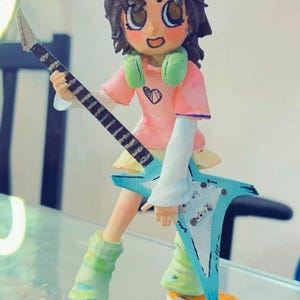 Puede incluir: Figura hecha a mano de una persona sosteniendo una guitarra en forma de estrella. La figura tiene cabello castaño, una camisa rosa con un diseño de corazón y auriculares verdes. La guitarra es azul claro con detalles en blanco y negro. La figura lleva ropa verde y amarilla.