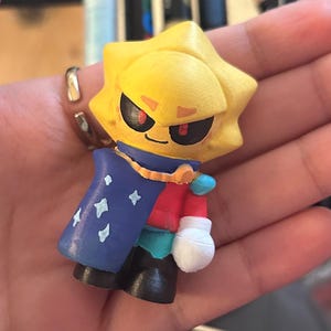 May include: A small, hand-painted figurine of a sun-like character with a stern expression. The figure wears a blue cape with white stars, a red shirt, and black boots. The character has a yellow head and a blue collar.