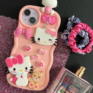 May include: A pink phone case with Hello Kitty and bow details, a perfume bottle, and two scrunchies. The phone case has a 3D design with Hello Kitty figures and paw prints. The perfume bottle has a floral design and gold accents.