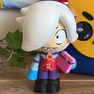 Op de afbeelding: Een beeldje van een stripfiguur met wit haar, een rood vest en een roze boek. Het personage heeft een lichte huidskleur, een brede glimlach en draagt een blauw en paars outfit. Het beeldje staat op een houten oppervlak.