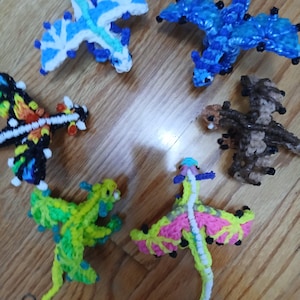 May include: A collection of handmade, colorful dragon figurines. The dragons are crafted from various materials, featuring shades of blue, green, yellow, pink, brown, and black. Each dragon has unique wing and body designs, with some including small, decorative details.