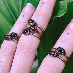 May include: A hand wearing multiple copper rings with red gemstone accents. The rings are stacked on two fingers.