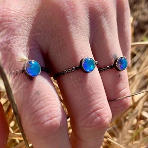 May include: Three rings with iridescent blue-green stones are displayed. The rings have a dark metal band. The stones are round and set in a simple bezel. The rings are on a hand with a neutral background.