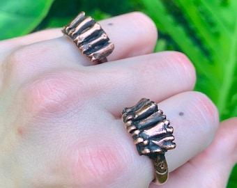 May include: Two bronze-colored rings, each featuring a textured design, are displayed on a hand. The rings have a unique, organic shape, and the metal has a slightly aged appearance. The background is a lush green with large leaves.