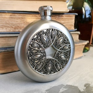 May include: A silver-colored, round flask with a decorative, ornate design on the front. The design is a detailed pattern of swirling shapes and leaves. The flask has a screw-on cap and is likely made of metal.