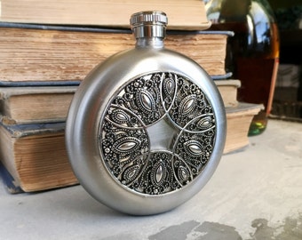 May include: A silver-colored, round flask with a decorative, ornate design on the front. The design is a detailed pattern of swirling shapes and leaves. The flask has a screw-on cap and is likely made of metal.