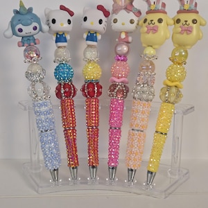 Puede incluir: Seis bolígrafos decorativos con figuras en la parte superior, incluyendo Hello Kitty. Los cuerpos de los bolígrafos están adornados con brillantes pedrería en varios colores: azul, rojo, rosa, naranja y amarillo. Los bolígrafos se exhiben en un soporte de plástico transparente.