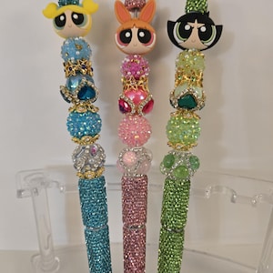 May include: Three Powerpuff Girls-themed pens. Each pen features a character's head at the top, with a body covered in sparkling rhinestones. The pens are in blue, pink, and green, with decorative jewel accents.