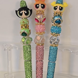 Puede incluir: Tres bolígrafos decorativos con adornos de personajes de dibujos animados. Los bolígrafos tienen barriles con incrustaciones de joyas en verde, rosa y azul. Cada bolígrafo tiene un diseño de personaje diferente, con detalles de joyas a juego y una parte superior brillante.