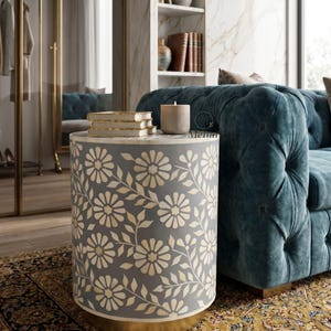 May include: A cylindrical side table featuring a grey base with a white floral and vine inlay pattern. The top surface holds a stack of three books and a beige cylindrical candle. The table has a solid, flat base and a smooth, rounded exterior.