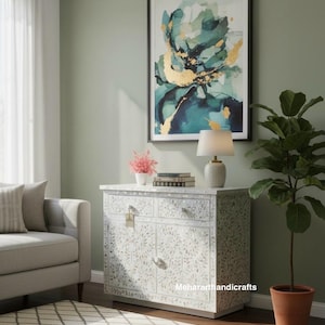 May include: A living room scene featuring a white cabinet with floral inlay, topped with a lamp and books. Above, a framed abstract painting in teal and gold hangs on a sage green wall. A sofa and patterned rug complete the decor.