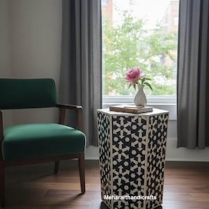 May include: A decorative side table with a geometric black and white pattern. A small white vase with a pink flower sits on top of the table. A green chair is to the left of the table. The table is approximately 20 inches tall.