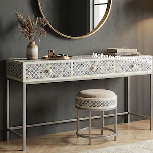 Puede incluir: Una mesa de tocador blanca con tres cajones, un taburete a juego y un espejo con marco dorado. La mesa y el taburete presentan un patrón decorativo. Un jarrón con flores secas y una bandeja con objetos pequeños están sobre la mesa. El texto "Mehararthhandicrafts" es visible.