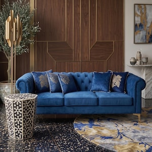 May include: A deep blue velvet tufted sofa with rolled arms and gold-toned legs, featuring five square throw pillows with gold embroidered floral patterns. The sofa is positioned in a room with dark terrazzo flooring and a wood-paneled wall.