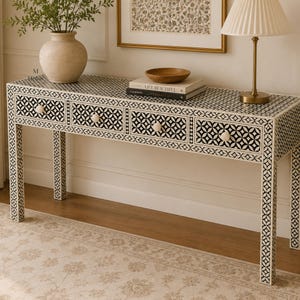 May include: A rectangular console table featuring a black and white geometric bone inlay pattern across its surface and four drawers with round white knobs. The table stands on four straight legs and is decorated with a vase and books.