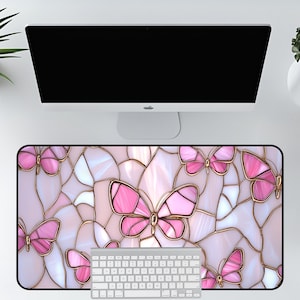 May include: A rectangular desk mat featuring a stained-glass butterfly design in shades of pink, white, and gold. The mat is placed under a computer monitor and keyboard, creating a decorative and functional workspace accessory.