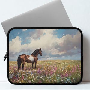 May include: Laptop sleeve featuring a painting of a brown and white horse standing in a field of wildflowers under a cloudy sky. The sleeve has a black zipper and a black border, with a gray laptop partially visible.