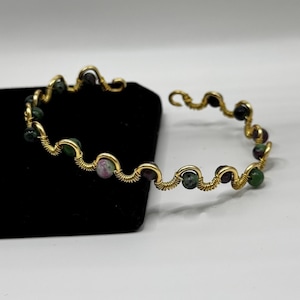 May include: A gold-colored wire-wrapped bracelet with alternating green, red, and black gemstone beads. The bracelet is resting on a black surface against a white background. The wire is intricately woven.