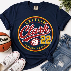 May include: A navy blue t-shirt featuring a circular graphic with the text CAITLIN CLARK, the number 22, and INDIANA FEVER in red, yellow, and white. The design includes a stylized basketball graphic centered within the circular emblem.